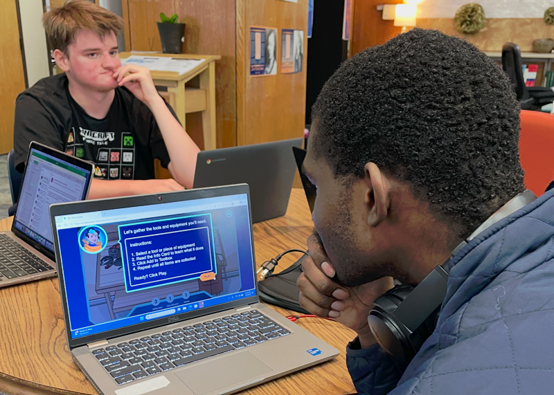 Two students sit at a table in a library, each looking at a laptop. One student's screen displays an educational game.